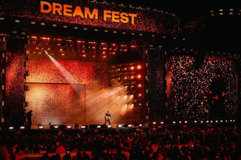 This image captures the energetic atmosphere of a live music festival at night. The main focus is the large, dynamic stage setup. Male artist, No Mercy’s Marty Cintron, standing in the center, plays a guitar under a bright spotlight. The stage is visually striking, with large screens on either side and a top banner all displaying a vibrant, fiery red and orange light pattern. The event's name, "DREAM FEST," is prominently featured in large, lit-up letters at the top of the stage. A diverse and large audience fills the foreground, their faces and hands visible as they watch the performance. The overall mood is one of excitement and a shared concert experience.