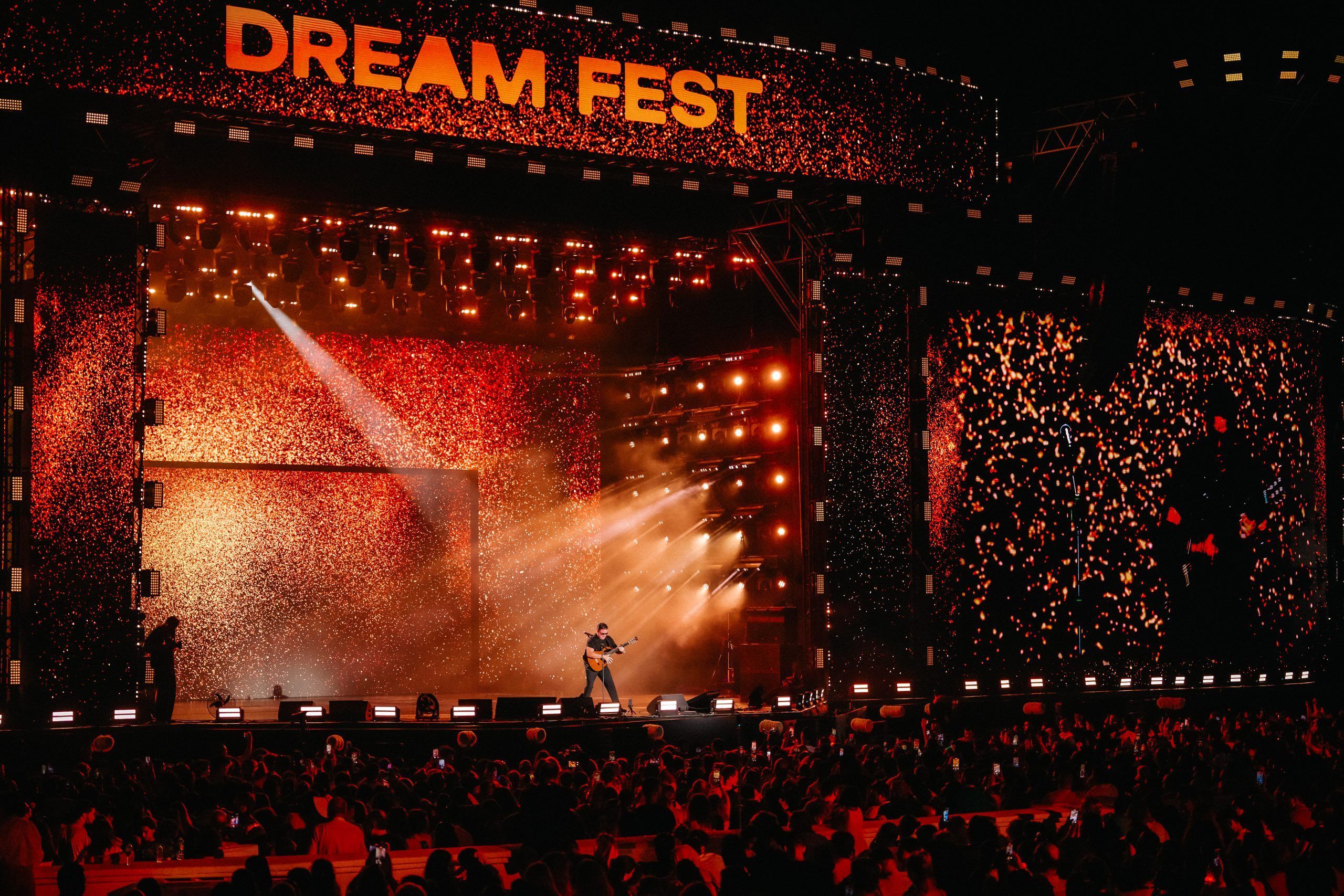 This image captures the energetic atmosphere of a live music festival at night. The main focus is the large, dynamic stage setup. Male artist, No Mercy’s Marty Cintron, standing in the center, plays a guitar under a bright spotlight. The stage is visually striking, with large screens on either side and a top banner all displaying a vibrant, fiery red and orange light pattern. The event's name, "DREAM FEST," is prominently featured in large, lit-up letters at the top of the stage. A diverse and large audience fills the foreground, their faces and hands visible as they watch the performance. The overall mood is one of excitement and a shared concert experience.