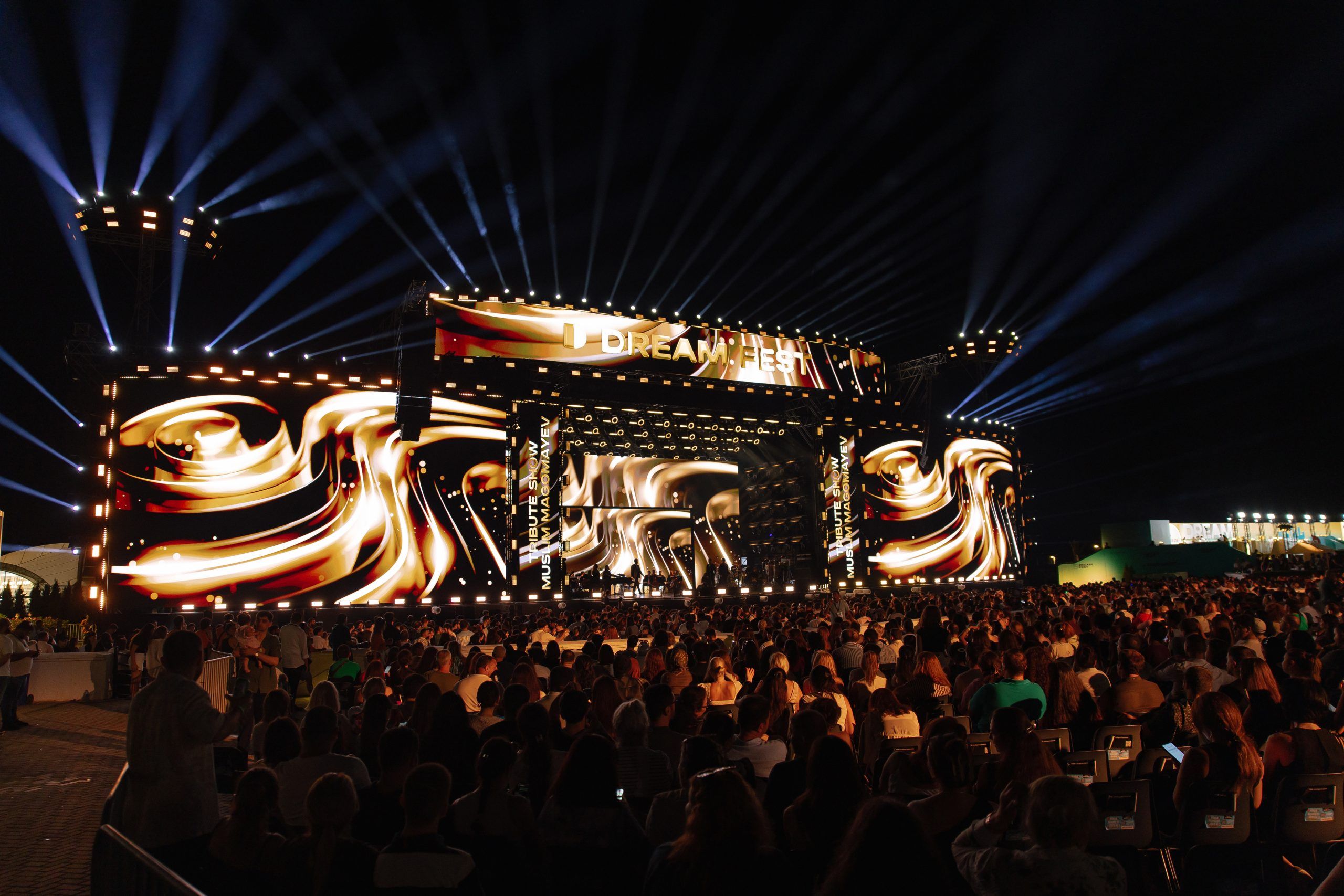 A massive outdoor stage at night with "DREAM FEST" written on the top. The stage is lit by spotlights and large screens displaying a swirling gold and brown pattern.