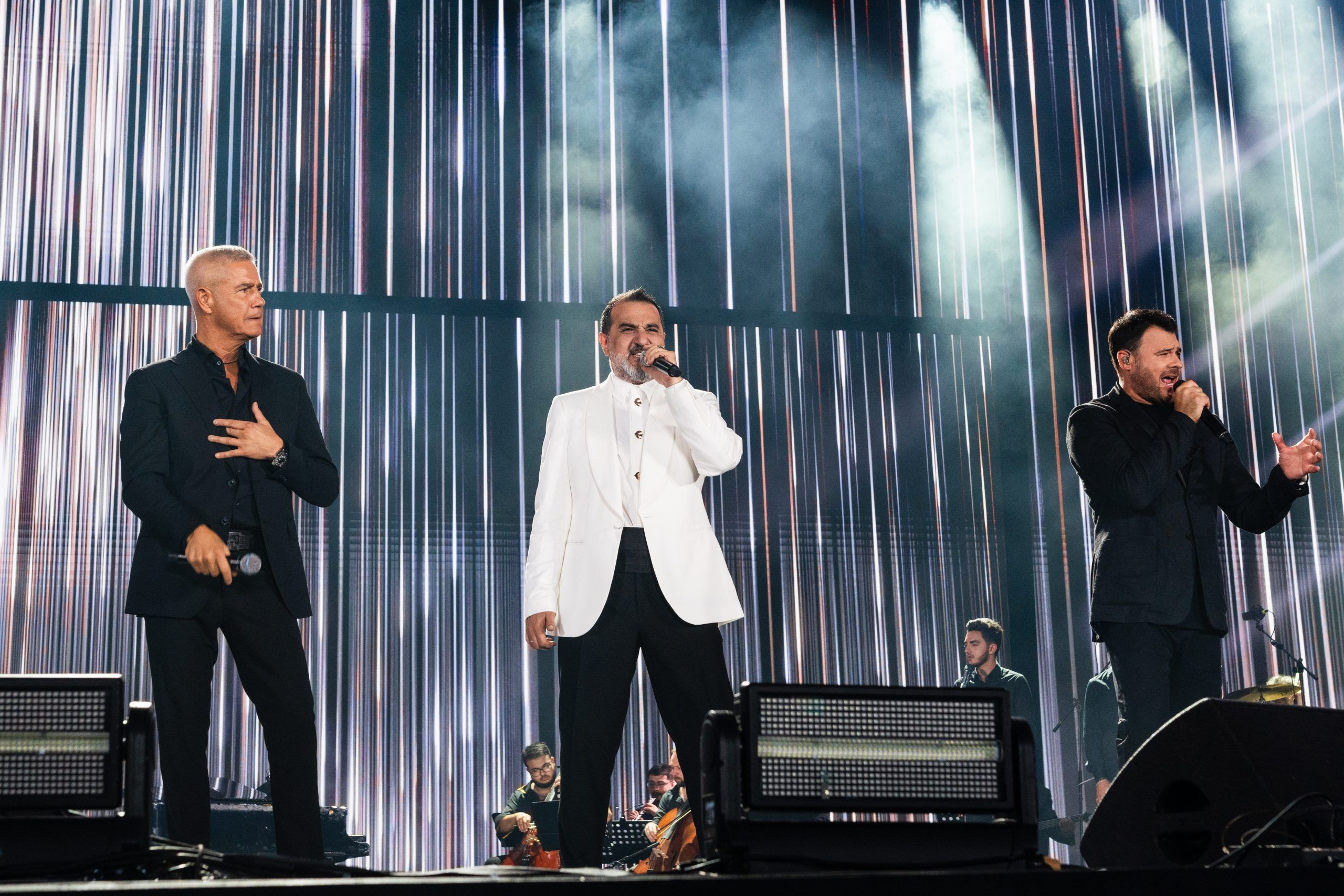 Three male singers in formal attire, two in black and one in a white jacket, stand on a stage with a background of vertical silver light beams.