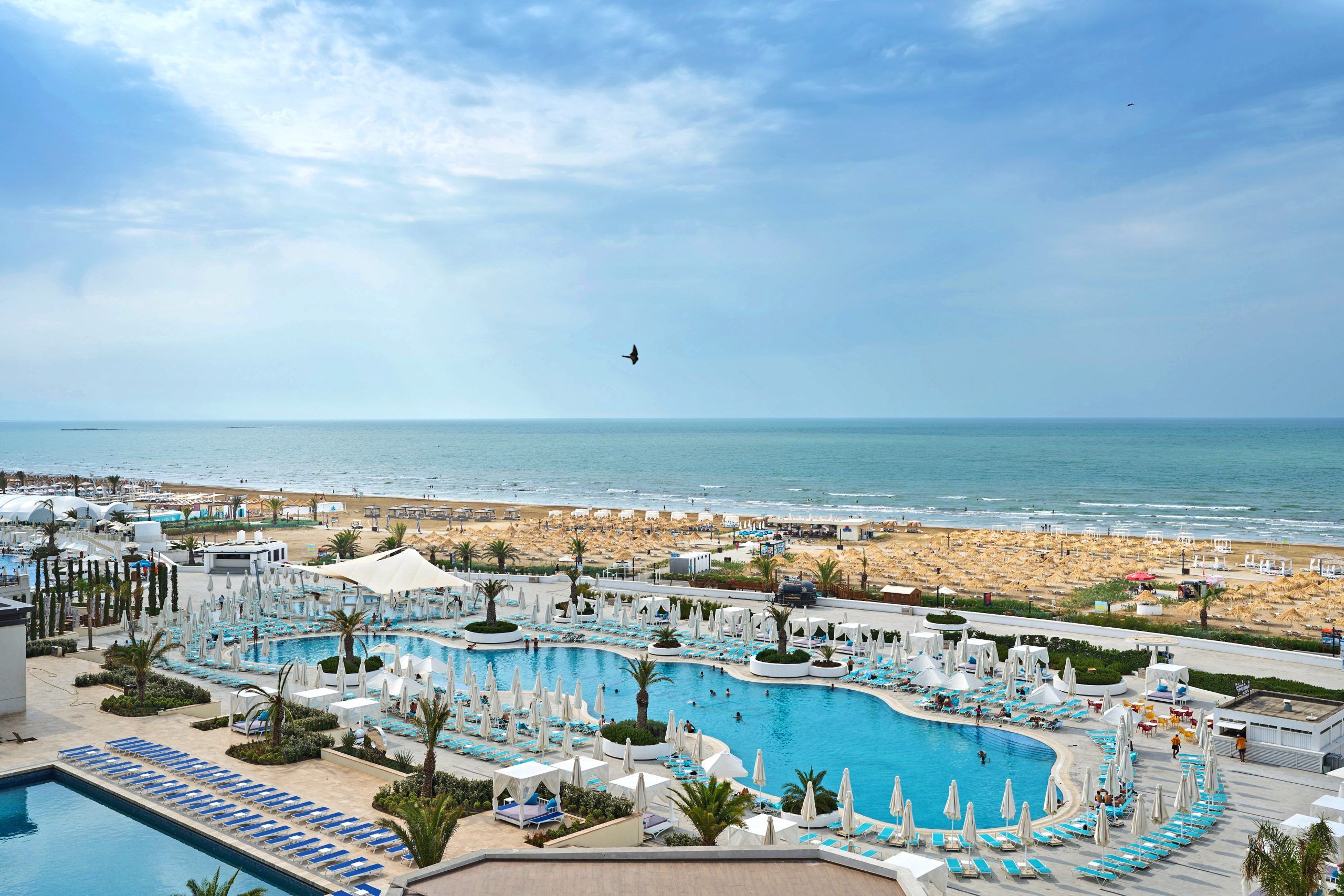 An elevated view of the Sea Breeze beach resort in Baku, Azerbaijan with a large, bright blue swimming pool, poolside lounges, and private cabanas. The scene overlooks the expansive beach and Caspian Sea under a blue sky.