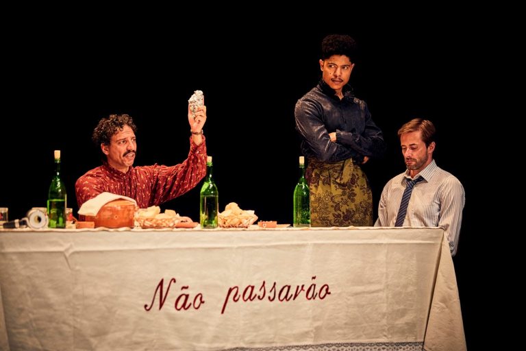 Three performers gathered around the embroidered dinner table in Catarina and the Beauty of Killing Fascists