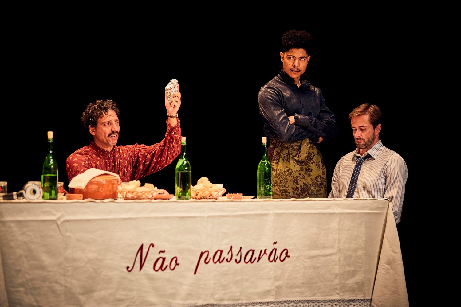 Three performers gathered around the embroidered dinner table in Catarina and the Beauty of Killing Fascists